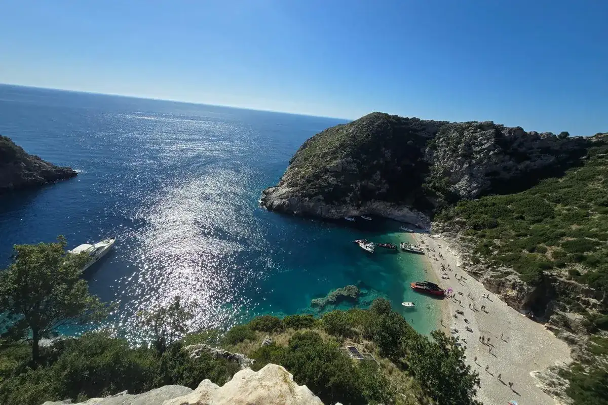 Grama Bay from the cliff top in afternoon sun: the headland, boats at anchor near the long pale beach and the open sea glittering beyond