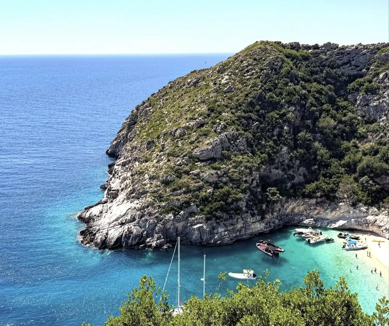 The green, rocky headland above Grama Bay seen from the hillside, with a sailing yacht and motorboats anchored off the pale beach and the open sea beyond