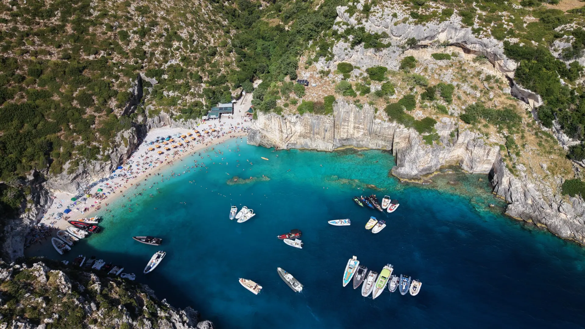 Grama Bay from above: a curved white pebble beach with sun umbrellas at the head of the cove, pale cliffs to the right and small boats at anchor in turquoise water